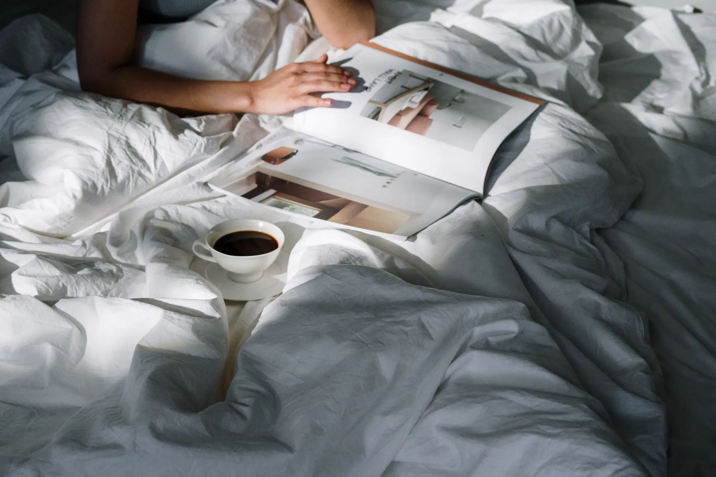 Person reading magazine in bed with coffee cup on white bedding